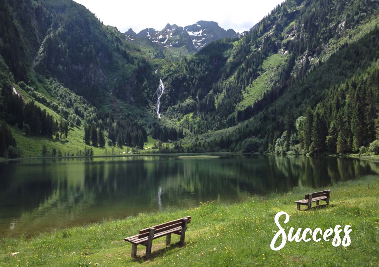 Photo of a mountain lake with a green meadow and two benches by the lake. The word “Success” at the bottom right.